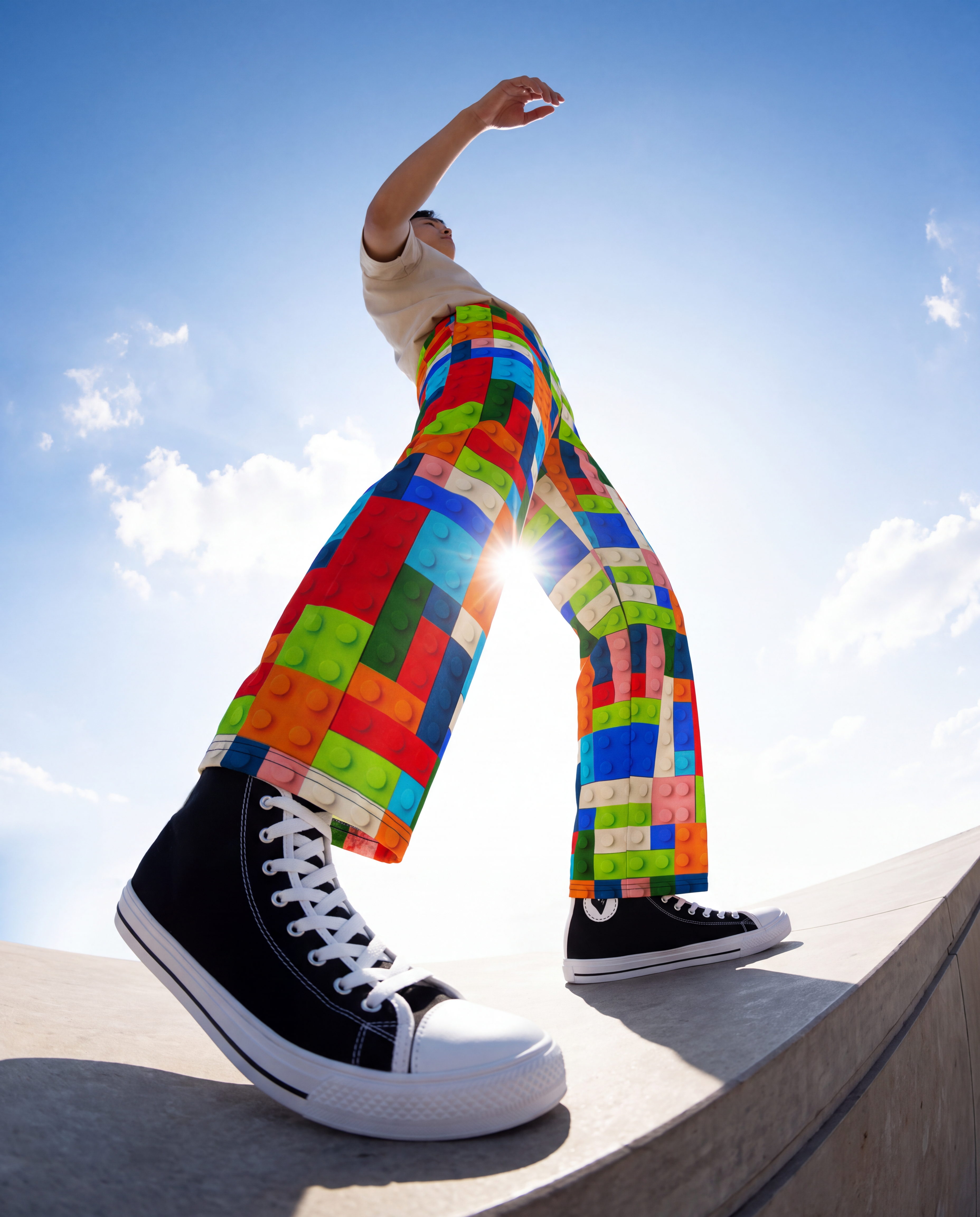 A person outdoors wearing bright multicolor pants with a block-studio pattern, black high-top sneakers, and a white top, standing on a concrete ledge against a blue sky.
