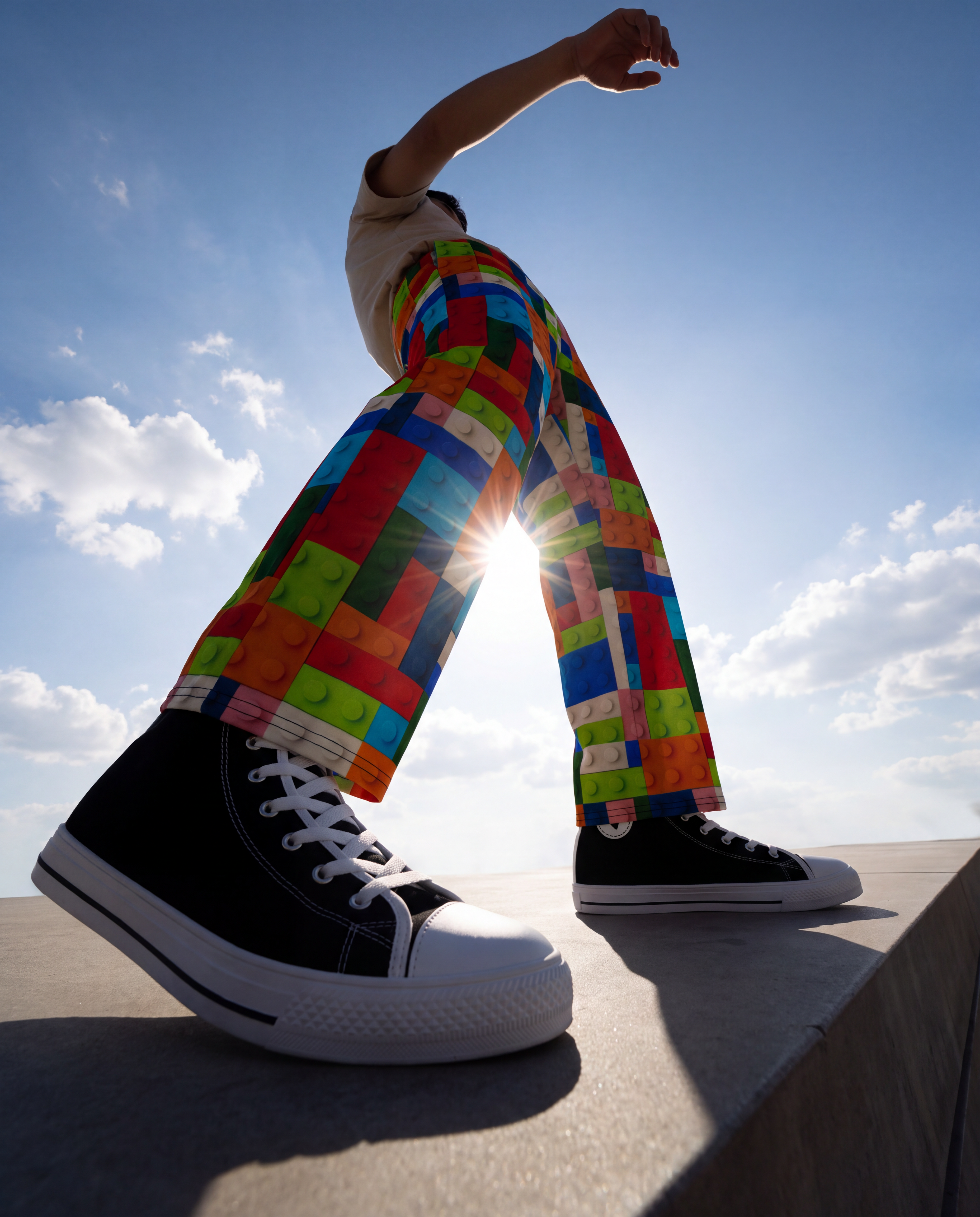 A person wearing colorful block-print pants and black high-top sneakers stands on a concrete ledge with the sun shining behind them against a blue sky.
