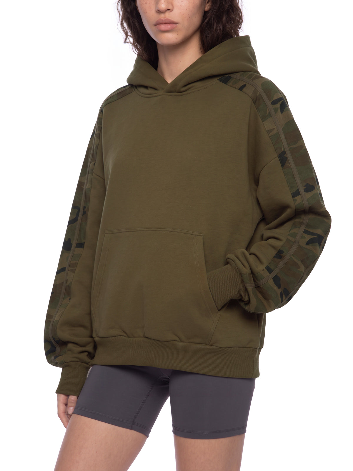 An oversized olive green pullover hoodie with camouflage-patterned sleeve panels, worn by a model with dark gray bike shorts against a plain white background.