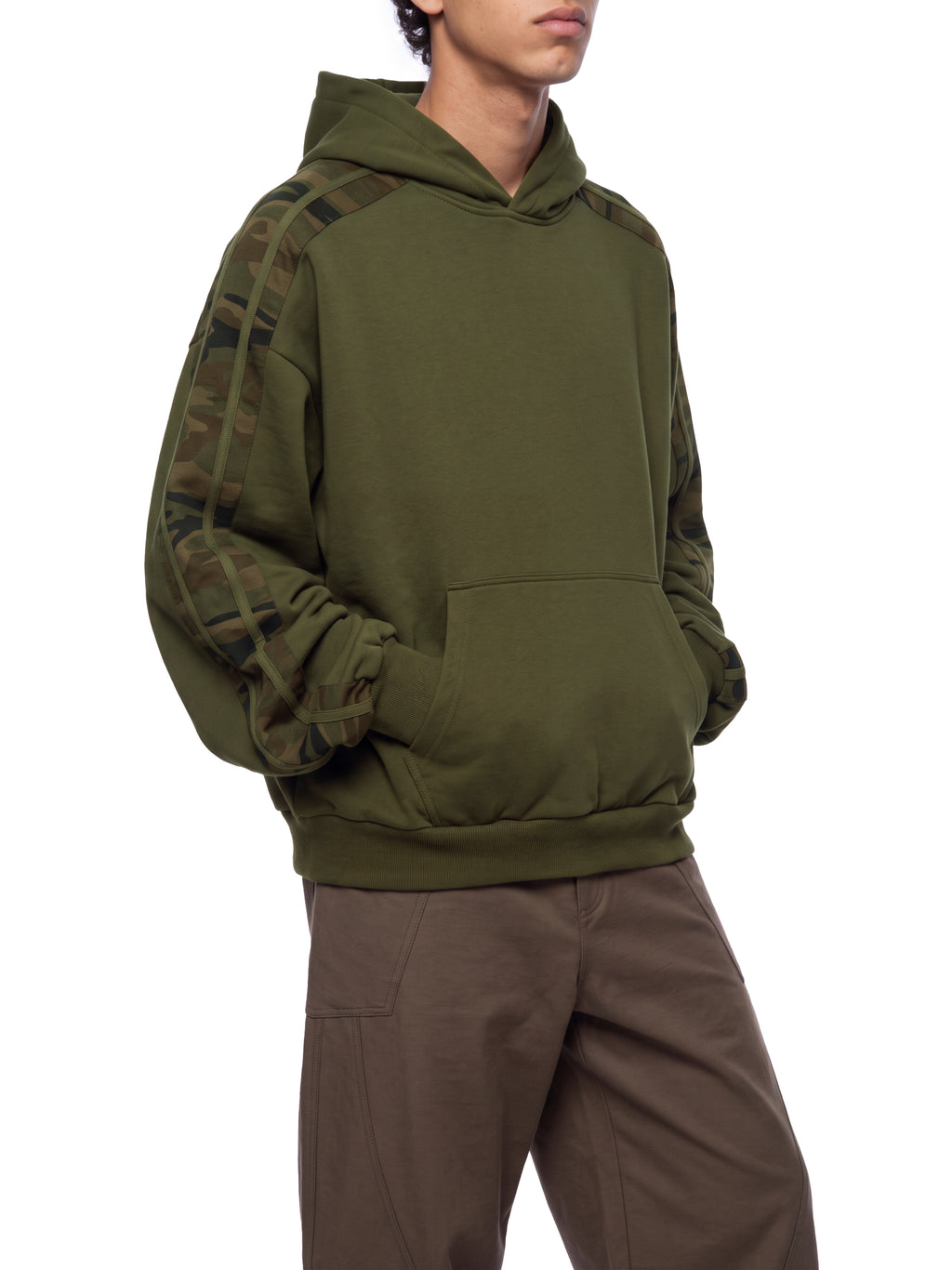 A person wearing an olive green pullover hoodie with camouflage-patterned sleeves, paired with brown pants, shown against a plain white background.