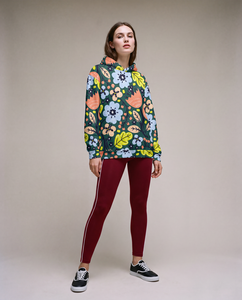 A woman modelling a dark green pullover hoodie with a vibrant multicolour floral pattern, paired with burgundy leggings and black sneakers, against a neutral beige studio background.