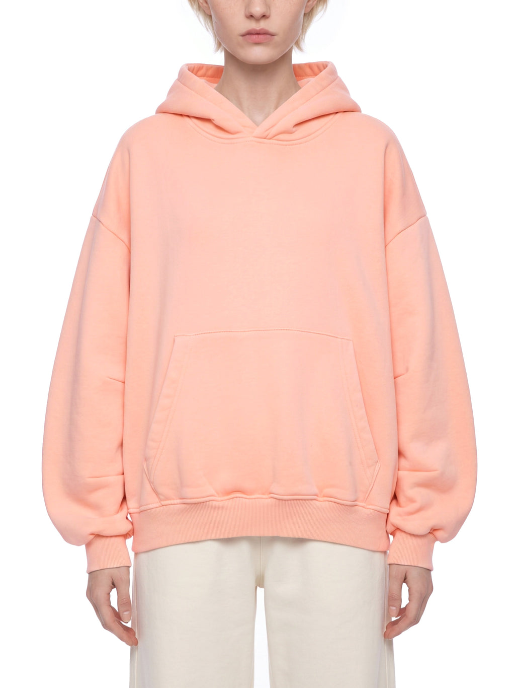 A model wearing an oversized peach-colored pullover hoodie with a kangaroo pocket, paired with cream-colored pants, shot against a solid white background.