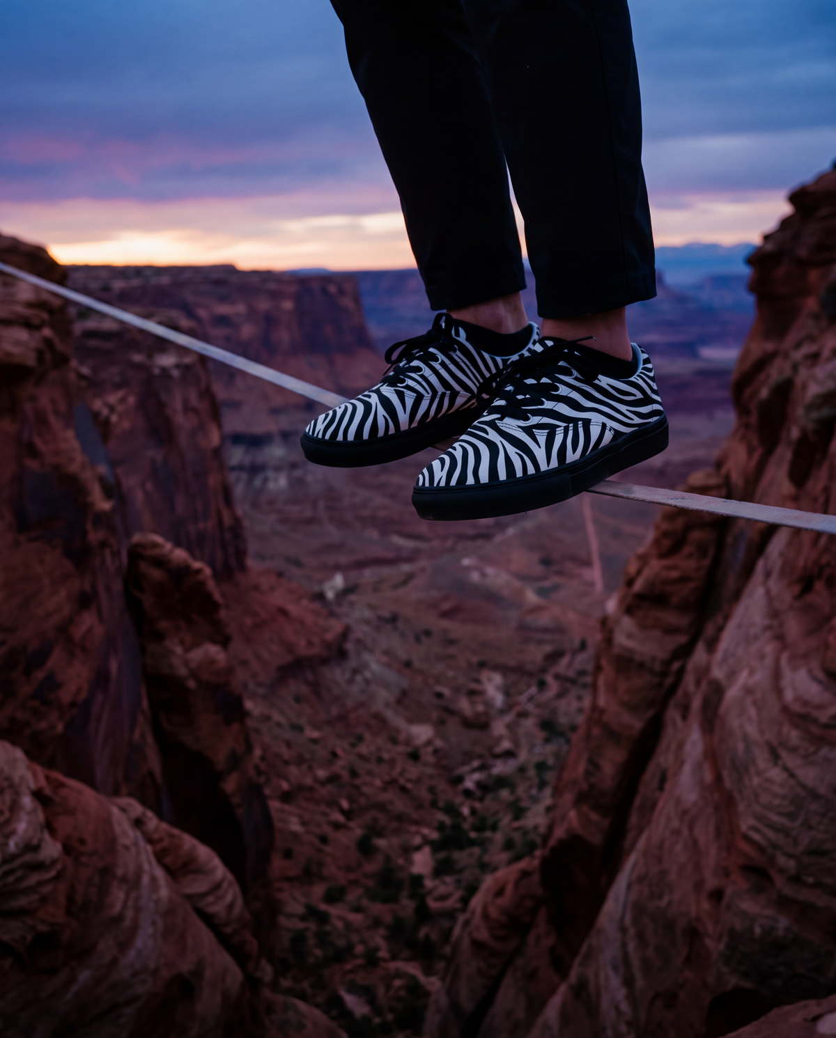 A person balancing on a tightrope over a deep canyon at sunset, wearing black-and-white zebra print sneakers and dark pants.