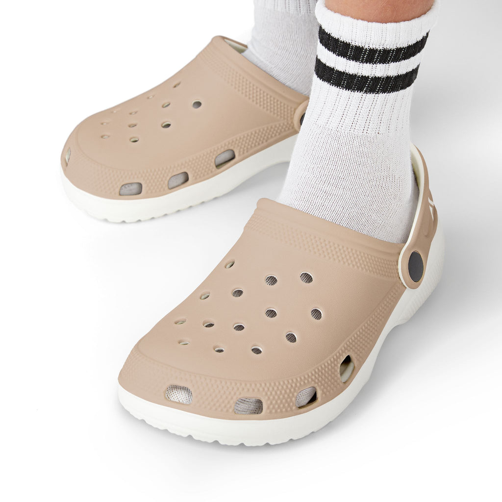 Beige Croc-style clogs with white soles, shown on a person's feet wearing white socks with black stripes, against a clean white background.